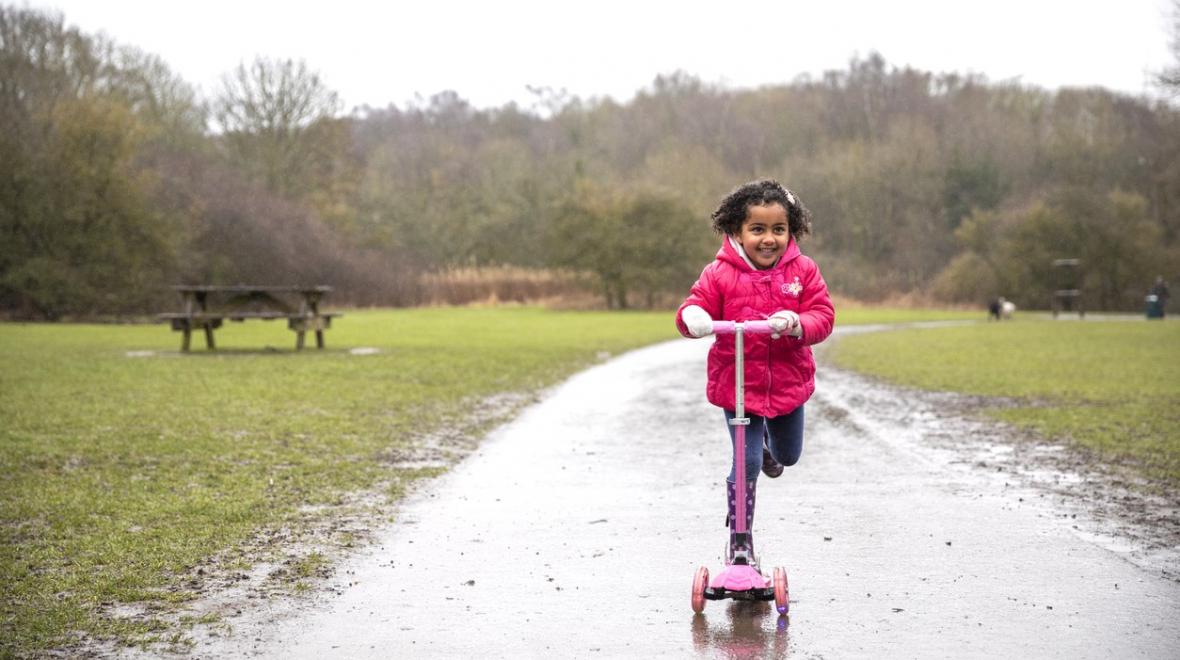 A girl in a pink jacket rides a scooter along a path through a park on a rainy day. She is smiling to be in best Seattle parks and playgrounds for rainy-day play